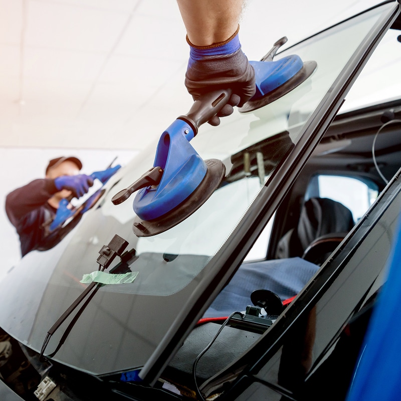 Technicien utilisant une ventouse double bleue pour installer ou remplacer un pare-brise de voiture dans un atelier.